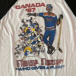 K&ouml;nnte beinhalten: Ein wei&szlig;es und schwarzes Baseball-T-Shirt mit einem Grafikdruck eines Hockeyspielers, der einen Hockeyschl&auml;ger h&auml;lt. Die Grafik enth&auml;lt den Text "CANADA '87" und "999". Das T-Shirt enth&auml;lt auch eine Liste von St&auml;dten und Daten f&uuml;r ein Hockeyturnier. Der Text "WHO GIVES A PUCK" ist auf der Unterseite des Shirts aufgedruckt.