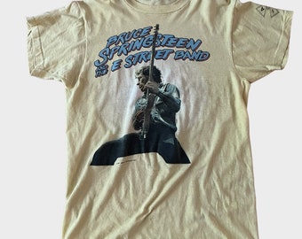 May include: Cream-colored t-shirt featuring a graphic of Bruce Springsteen playing guitar, with the text "Bruce Springsteen and the E Street Band" in blue and red. The shirt has a vintage look.