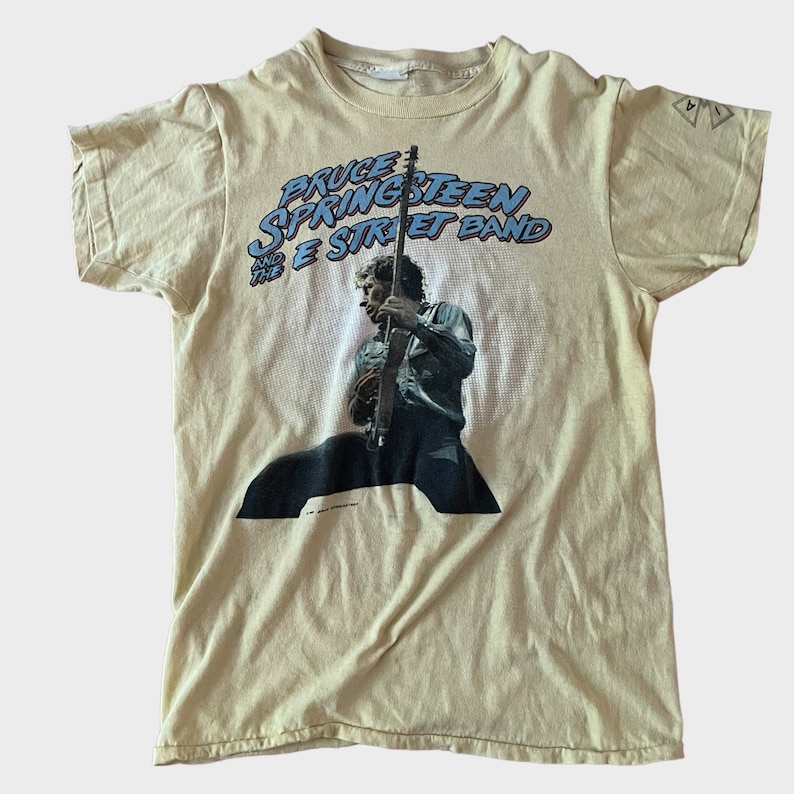 May include: Cream-colored t-shirt featuring a graphic of Bruce Springsteen playing guitar, with the text "Bruce Springsteen and the E Street Band" in blue and red. The shirt has a vintage look.