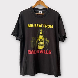 Puede incluir: Camiseta negra con un estampado gráfico amarillo y rojo de una mujer tocando la batería. El texto "BIG BEAT FROM" y "BADSVILLE" está impreso en la camiseta.