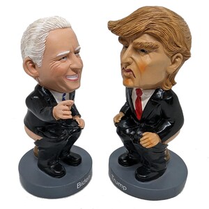 May include: Two ceramic figurines of political figures, one with a white bald head and the other with a blonde hairstyle, both wearing black suits and red ties. The figurines are sitting on a toilet, with the names "Biden" and "Trump" written on the bases.