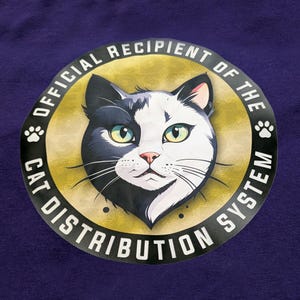 Peut inclure: T-shirt violet avec un motif circulaire représentant un chat noir et blanc aux yeux verts et au nez rose. Le motif est entouré d'un texte blanc qui dit "OFFICIAL RECIPIENT OF THE CAT DISTRIBUTION SYSTEM". Des empreintes de pattes sont de chaque côté.