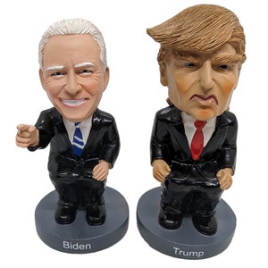 May include: Two bobblehead figurines of political figures. The figurine on the left is of Joe Biden, wearing a black suit and blue tie, with a smiling expression and his left hand pointing forward. The figurine on the right is of Donald Trump, wearing a black suit and red tie, with a frowning expression. Both figurines are sitting on gray bases with their names printed below.