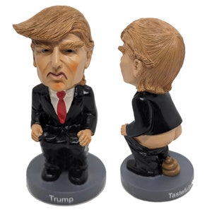 May include: A figurine of a person in a black suit and red tie, with a brown hairstyle. The figurine is in a seated position with the text "Trump" on the base. The figurine is also shown from the back, with the text "Tastefully Offensive" on the base.