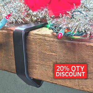 May include: A matte black metal stocking holder hook clamped onto a thick wooden mantelpiece. The hook features a smooth, curved design. A red rectangular graphic with white text reading 20% QTY DISCOUNT is visible on the wood surface.