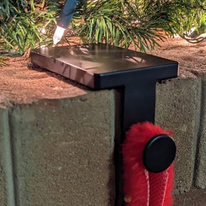 Può includere: Porta calze in metallo nero montato su un muro di mattoni. Il supporto ha una parte superiore piatta e un supporto verticale. Una calza rossa è appesa al supporto.