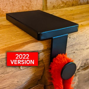May include: Black metal stocking holder with a flat top and a hook. The holder is mounted on a wooden surface. 2022 VERSION is written on the surface.
