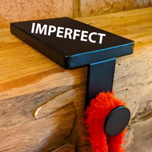 Op de afbeelding: Zwarte metalen kousenhouder met het woord "IMPERFECT" erop gedrukt. De houder is bevestigd aan een houten oppervlak en heeft een rode, pluche kous aan de haak.