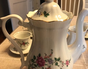 May include: White porcelain teapot with a floral design and gold trim. The teapot has a lid with a knob on top. The teapot is sitting on a table with a checkered tablecloth.