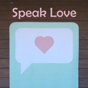 May include: A light blue and white quilt with a speech bubble design and a pink heart inside. The quilt has the text "Speak Love" at the top and "Slice of Pi Quilts" at the bottom.