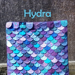 May include: A purple and blue quilt with a scale pattern. The quilt is made of fabric with a batik design. The quilt is labeled "Hydra" and "Slice of Pi Quilts".