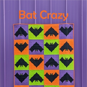 May include: A purple wall with a quilt hanging on it. The quilt is made of orange, green, and purple squares with black bats on each square. The quilt is titled "Bat Crazy" and has a black and white circle with the text "Slice of Pi Quilts" at the bottom.