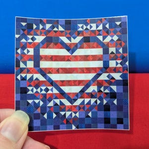 May include: A square sticker featuring a heart design with red and white stripes, surrounded by a mosaic of blue, red, and white geometric shapes. The sticker is held against a backdrop of blue and red.