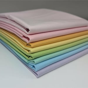 May include: A stack of seven folded fabric squares in a gradient of pastel colors. The colors include light pink, soft peach, pale yellow, light lime green, mint green, sky blue, and lavender. Each piece of fabric is neatly folded and stacked.