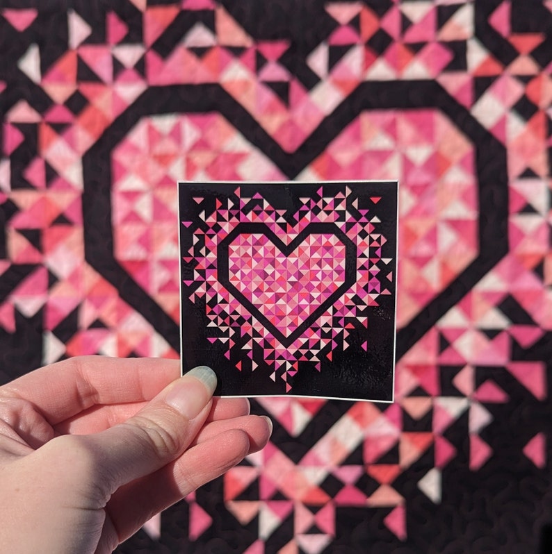 An all pink Mini Exploding Heart quilt hanging on a wall with a matching sticker being held in front