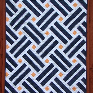 Peut inclure: Une courtepointe bleu marine et blanche avec un motif géométrique de rayures diagonales et de carrés orange. La courtepointe est faite avec un fond blanc et des rayures bleu marine qui créent un motif en losange. Les carrés orange sont répartis uniformément sur toute la courtepointe.