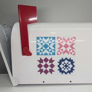 May include: White mailbox with a red flag and decorative quilt block designs in blue, pink, purple, and navy. The mailbox has a silver side panel and is on a white surface.
