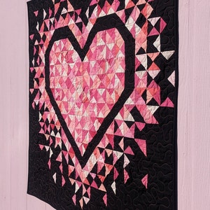 An all pink Mini Exploding Heart quilt with a black background fabric hanging on a light pink wall