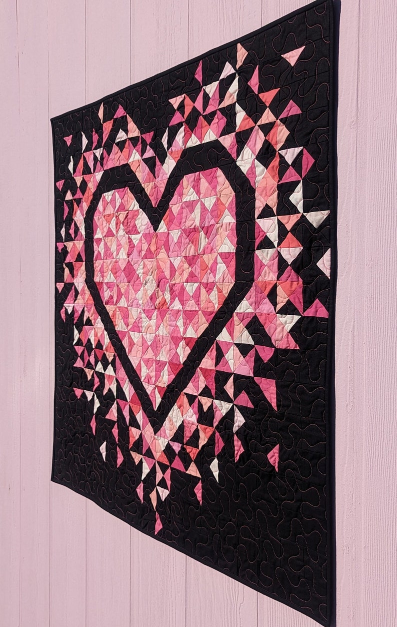 An all pink Mini Exploding Heart quilt with a black background fabric hanging on a light pink wall