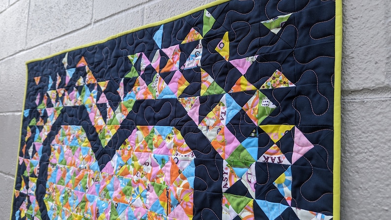 A colorful Mini Exploding Heart quilt made with a navy background fabric hanging on a wall