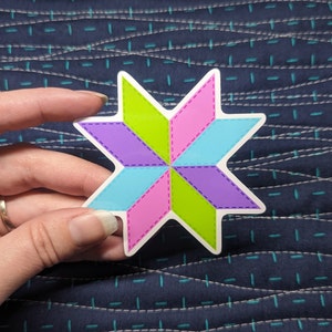 May include: A colorful star-shaped sticker with pink, green, blue, and purple sections. The sticker has a white background and a dashed line design around the edges.
