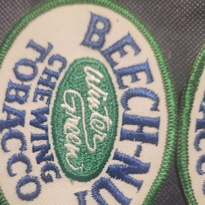 Könnte beinhalten: Ein weißer ovaler Patch mit grünem und blauem Besatz. Der Patch trägt den Text "Beech-Nut Chewing Tobacco" in blauer Schrift und ein grünes Oval mit dem Text "Blue for Flavor" im Inneren.