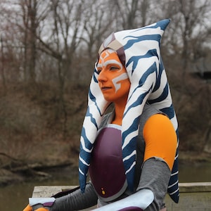 Può includere: Una donna con un copricapo a strisce blu e bianche con pittura per il viso arancione e bianca. Indossa un pezzo toracico viola e argento e un abito grigio e arancione.