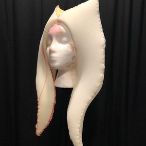 以下が含まれることがあります： 尖ったトップと長く流れるサイドを持つ白いフォームヘッドピース。ヘッドピースはピンクの糸で縫い合わされています。
