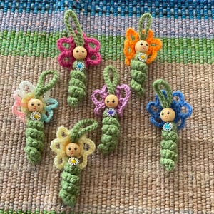May include: A selection of handmade crochet flower ornaments in various colours. Each ornament features a wooden bead head, a green body, and colourful flower petals. The flowers include pink, orange, blue, yellow, and pastel rainbow hues.