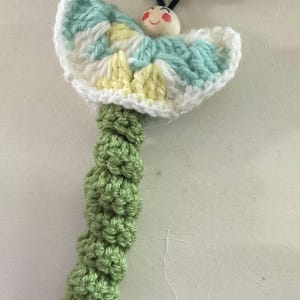 May include: A handmade crochet butterfly with a smiling wooden face, black antennae, and wooden bead tips. The butterfly's wings are white, blue, and yellow. The stem is green and textured.