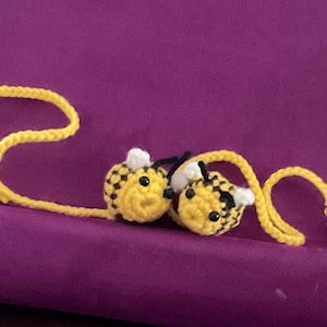 May include: Crocheted bee bookmarks with yellow and black striped bodies, white wings, and black eyes. The bees are connected by a yellow cord with beads and a tassel. The bookmarks rest on a purple surface.