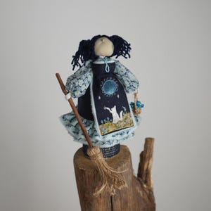 Könnte beinhalten: Handgefertigte Puppe in einem dunkelblauen Kleid mit einem Himmelsdesign, die einen Besen und einen kleinen Korb hält. Die Puppe hat dunkelblaues Garnhaar und hellblau gemusterte Ärmel und einen Rock. Die Puppe steht auf einem Holzsockel.