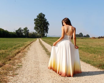 May include: A woman in a white and yellow ombre dress stands on a gravel path in a field. The dress has a fitted bodice and a flowing skirt. The path leads towards a large tree in the distance, with green fields on either side.