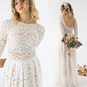 Puede incluir: Un vestido de novia de encaje blanco con mangas largas y espalda baja. El vestido tiene un corpiño ajustado y una falda fluida. La modelo lleva una corona de flores y sostiene un ramo de flores.