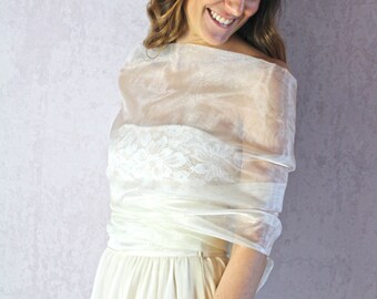 May include: A sheer, ivory-colored shawl draped over a cream-colored dress. The dress features a floral lace bodice. The shawl is made of a lightweight, flowing fabric, creating a soft, elegant look. The overall style suggests a formal or bridal theme.