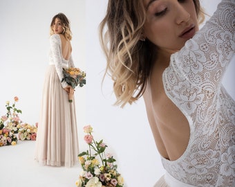 May include: A white lace wedding dress with long sleeves and a low back. The dress has a fitted bodice and a flowing skirt. The model is holding a bouquet of flowers.