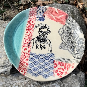 May include: A white ceramic plate with a blue and red design. The plate features a portrait of Ruth Bader Ginsburg with the text "Notorious RBG".