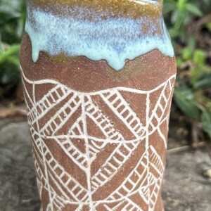 May include: A handmade ceramic vase with a unique design. The vase features a brown base with a white geometric pattern and a light blue and brown rim. The vase is approximately 5 inches tall.