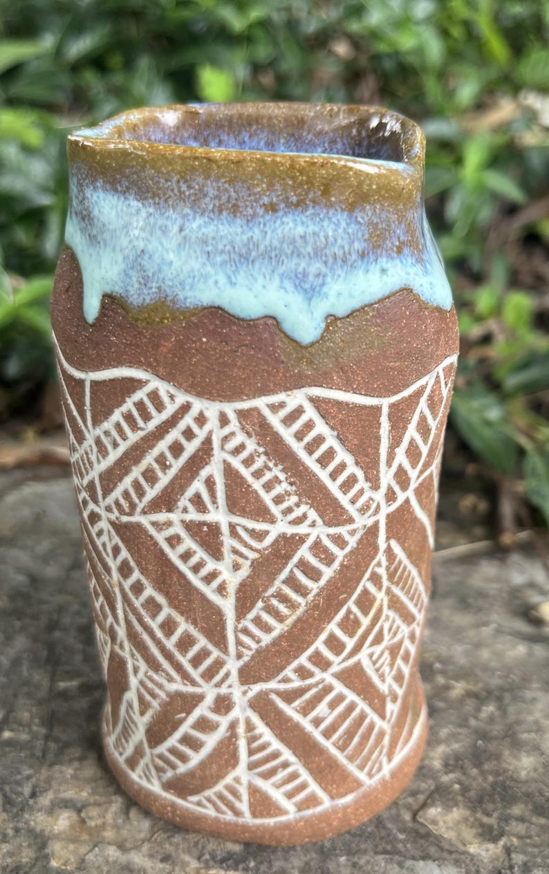 May include: A handmade ceramic vase with a unique design. The vase features a brown base with a white geometric pattern and a light blue and brown rim. The vase is approximately 5 inches tall.