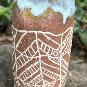 May include: A handmade ceramic tumbler with a brown body and a white geometric pattern. The rim is glazed with a blue and brown color. The tumbler is a unique piece of art.