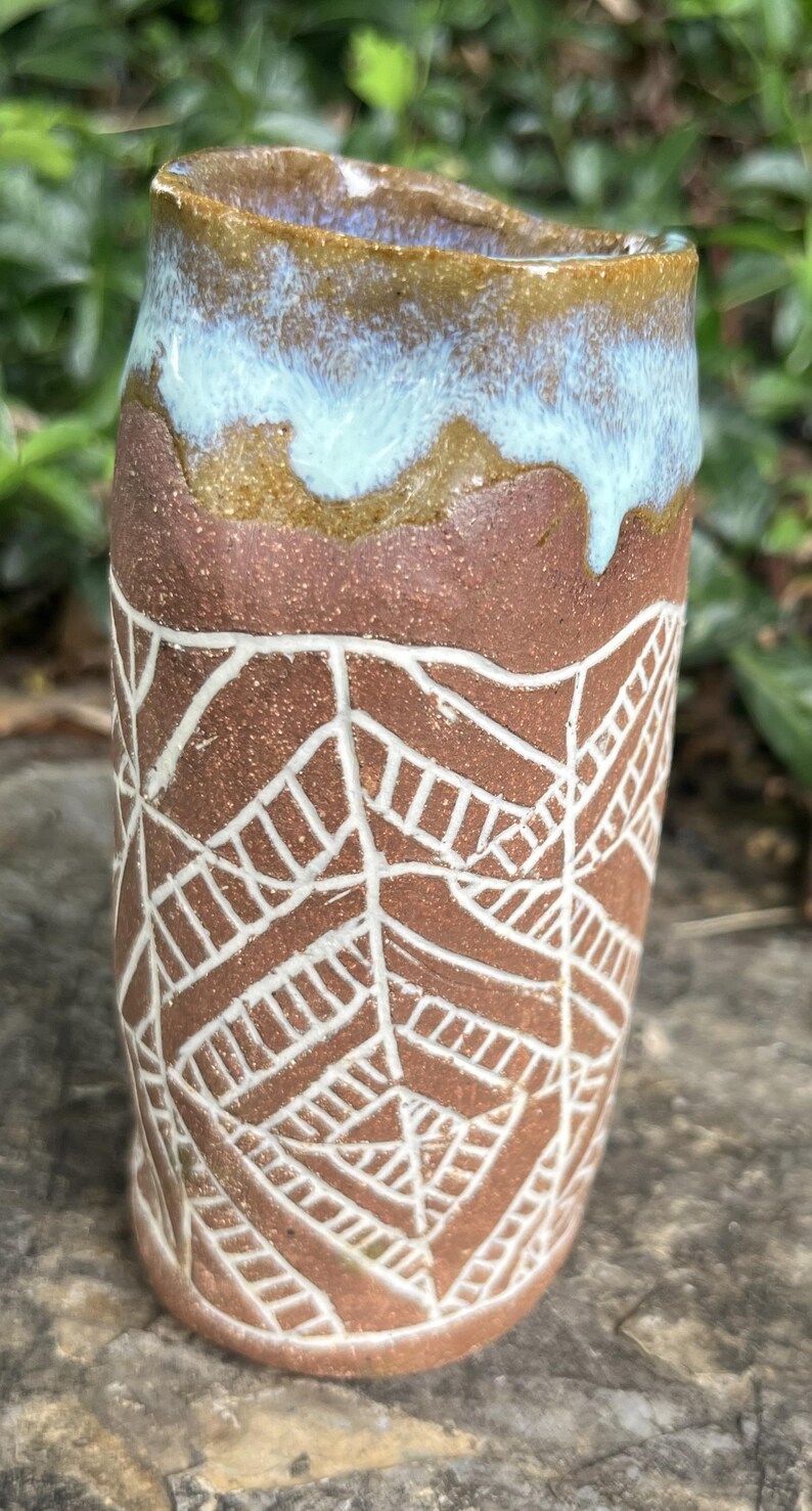 May include: A handmade ceramic tumbler with a brown body and a white geometric pattern. The rim is glazed with a blue and brown color. The tumbler is a unique piece of art.