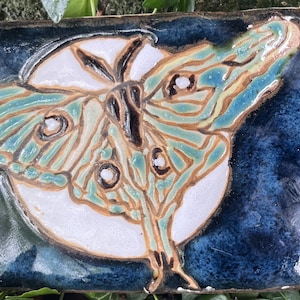 May include: Rectangular ceramic tile featuring a Luna moth design. The moth has teal and brown wings with a white body, set against a dark blue background. The tile is likely handmade and suitable for home decor.