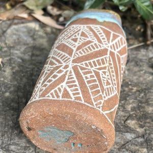 May include: A handmade ceramic tumbler with a brown base and a white geometric pattern. The rim is light blue, and the bottom has a turquoise design. The tumbler is resting on a gray stone surface.