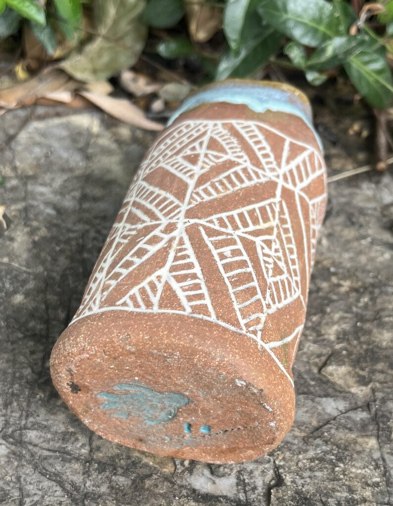 May include: A handmade ceramic tumbler with a brown base and a white geometric pattern. The rim is light blue, and the bottom has a turquoise design. The tumbler is resting on a gray stone surface.