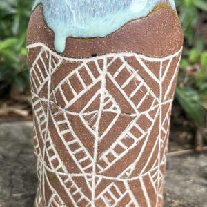 May include: Handmade ceramic tumbler with a brown base and a light blue glazed rim. The tumbler features a white geometric pattern etched into the brown clay. The design is a unique, handcrafted piece of pottery.