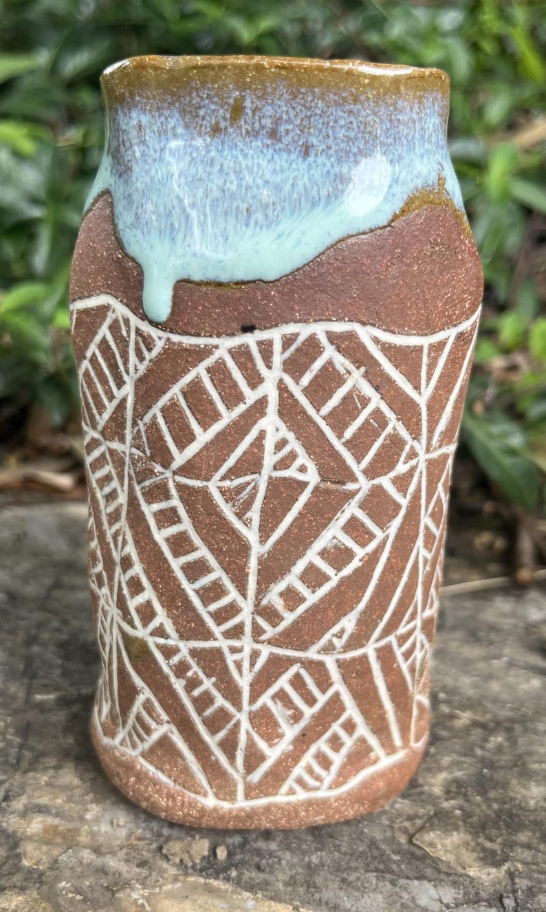 May include: Handmade ceramic tumbler with a brown base and a light blue glazed rim. The tumbler features a white geometric pattern etched into the brown clay. The design is a unique, handcrafted piece of pottery.