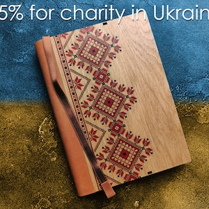 Puede incluir: Un diario de madera con una cubierta de cuero marrón y un diseño bordado en rojo y negro. El diario está sobre un fondo azul y dorado. El 15% de las ventas se donará a la caridad en Ucrania.