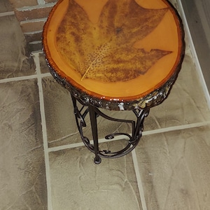May include: A small, round side table with a unique design. The tabletop is made from a cross-section of a tree trunk, with a large leaf design embedded in the amber-colored resin. The table has a wrought iron base.
