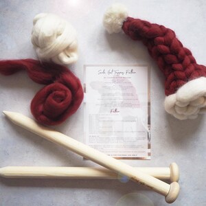May include: A red and white Santa hat topper knitting kit with instructions, two knitting needles, and red and white wool. The instructions are titled "Santa Hat Toppers Pattern by Laughlin Ashton Designs".