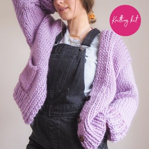 May include: A lilac knitted cardigan with a button closure and pockets. The cardigan is shown being worn over a grey t-shirt and black dungarees. The image includes the text "Knitting kit".